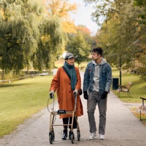 En äldre kvinna promenerar med rullator utomhus. Bredvid henne går en man. De pratar med varandra.