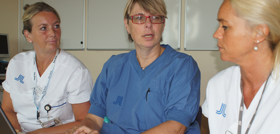 Halvbild på en kvinnlig läkare som sitter vid ett skrivbord med en sjuksköterska på var sida.