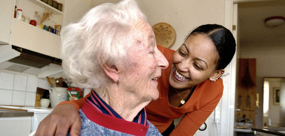 En kvinna ler och håller en äldre kvinna om axlarna. Köksmiljö.