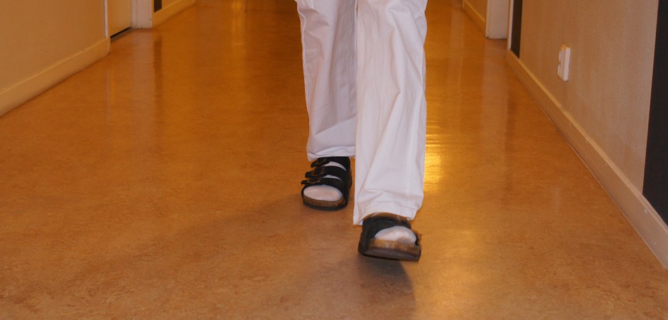 Ben i vita byxor och svarta sandaler går på ett brunt plastgolv.
