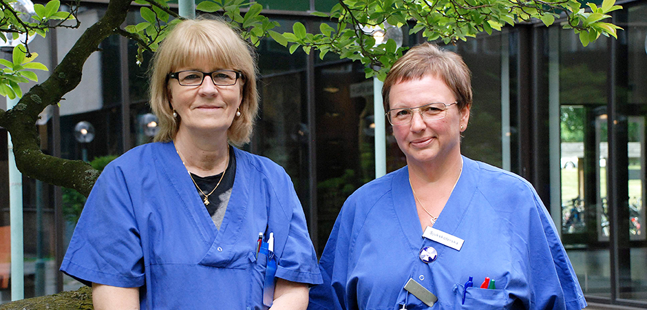 Två kvinnor i blåa sjukhuskläder står i utomhusmiljö.