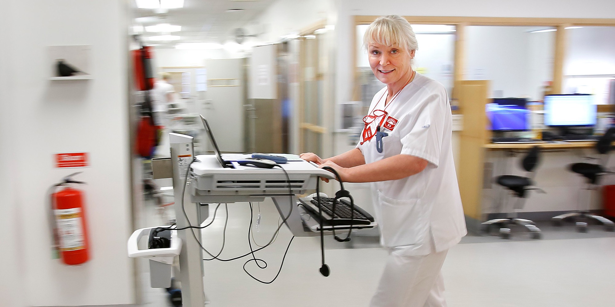 Läkaren Kerstin Skog Tigerström i sjukhuskläder står vid ett arbetsbord i en sjukhuskorridor.