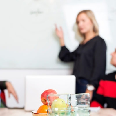 kvinna står vid whiteboard och tre personer sitter vid ett bord.