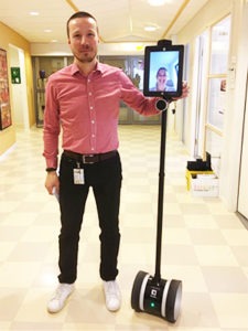 en man i en sjukhuskorrdior håller i en robot som består av en segway, samt en pinne med en ipad-skärm i ögonhöjd. 