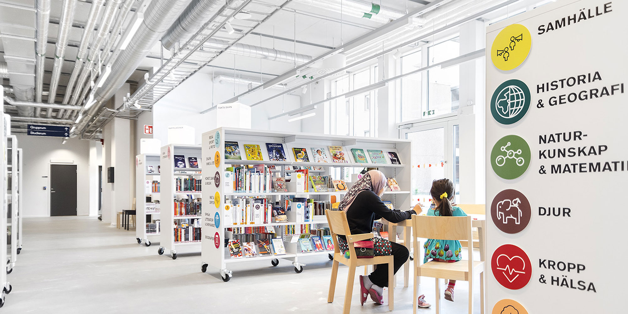 Interiör från bibliotek. vita hyllor, vitt tak, två personer sitter vid ett bord med ryggen mot kameran.