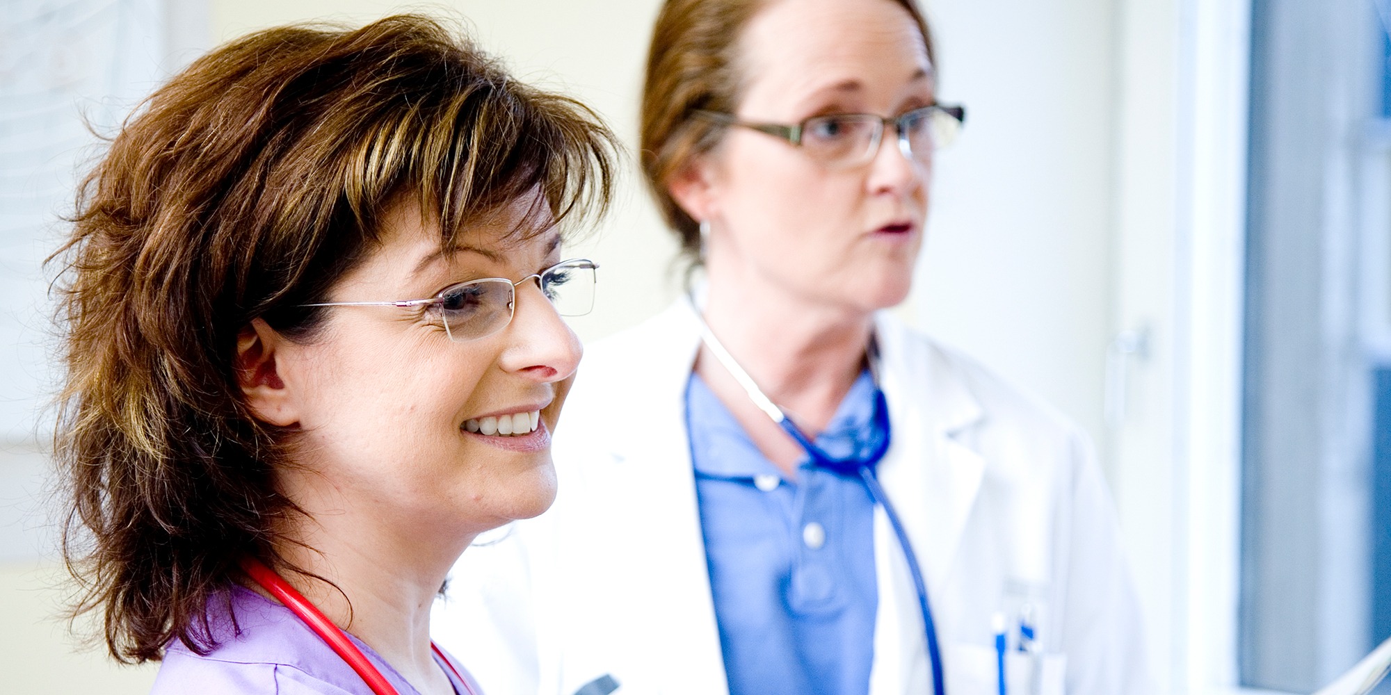 närbild på två kvinnliga läkare snett från sidan, i sjukhuskläder med stetoskop runt halsarna.