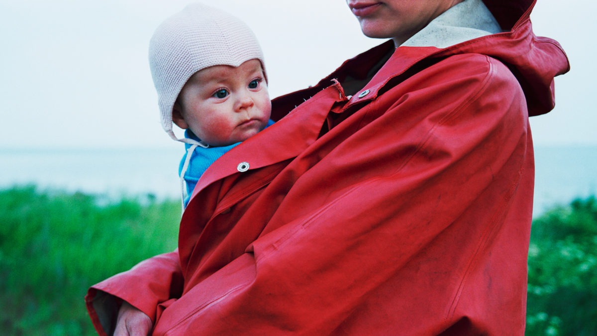 Person står framför hav iklädd röd regnjacka med bebis i famnen.