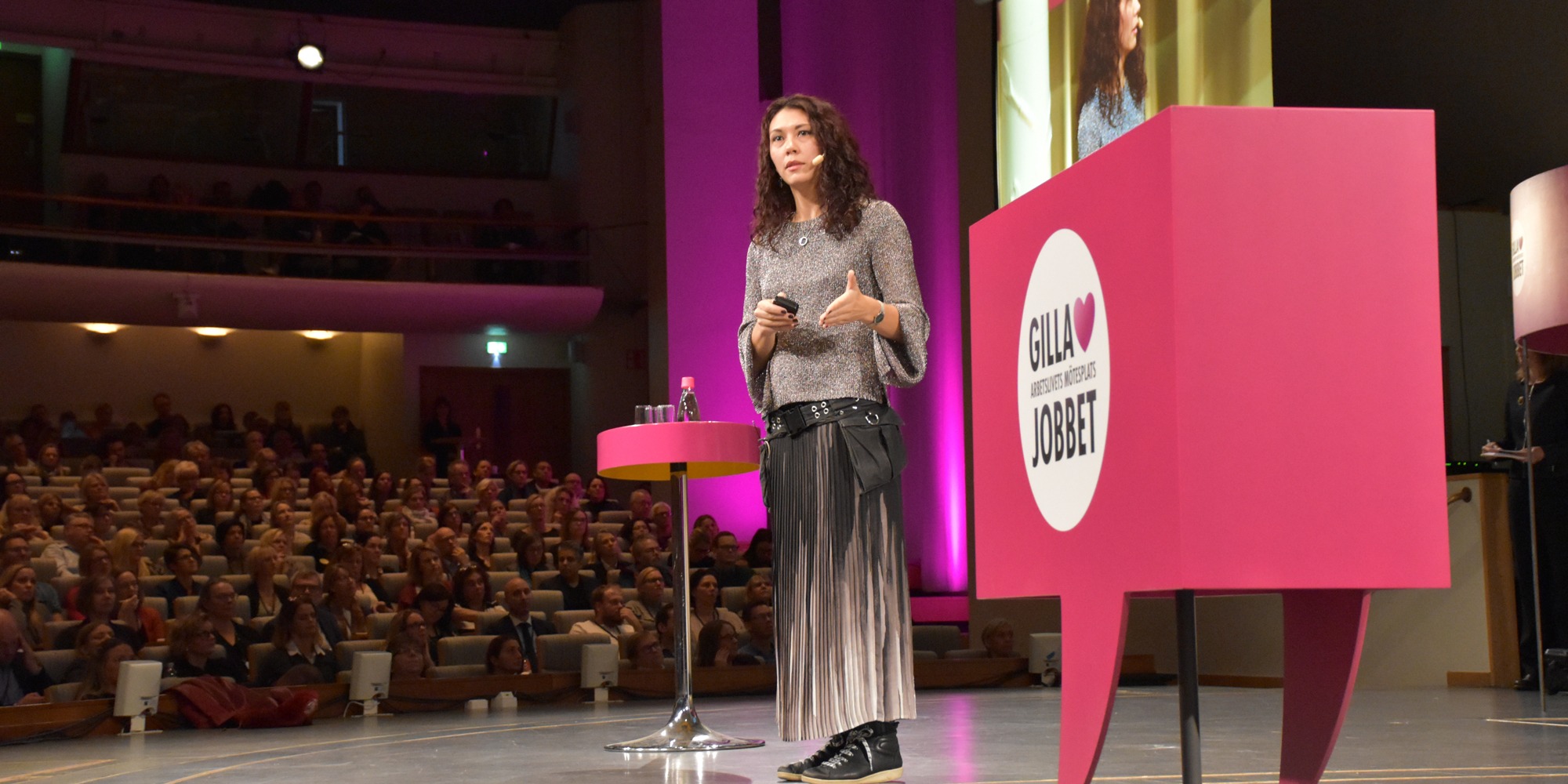 Forskaren Cecilia Berlin, klädd i lång kjol, talar till en stor publik från en scen på arbetsmiljökonferensen Gilla Jobbet