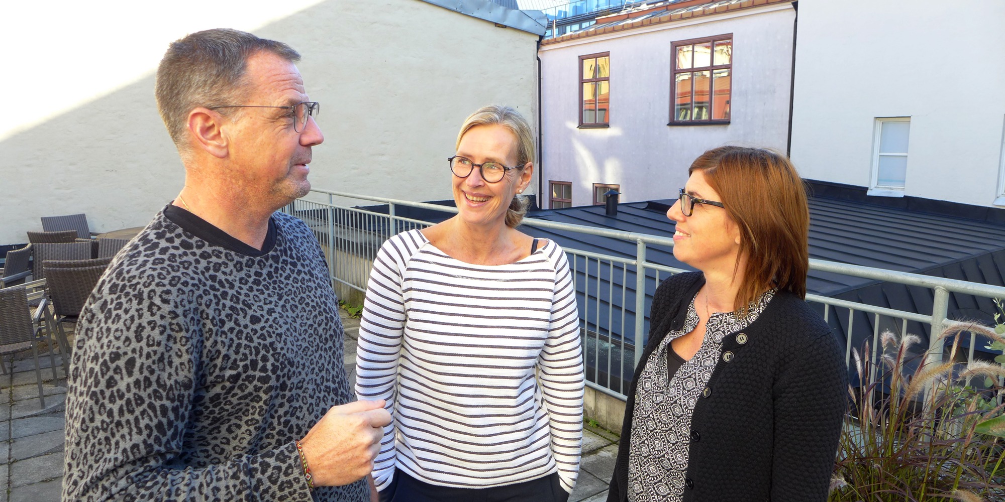 Johan Mellnäs, Petra Salino och Stina Ellerfelt Sköld samtalar leende med varandra på en terrass med husväggar i bakgrunden.