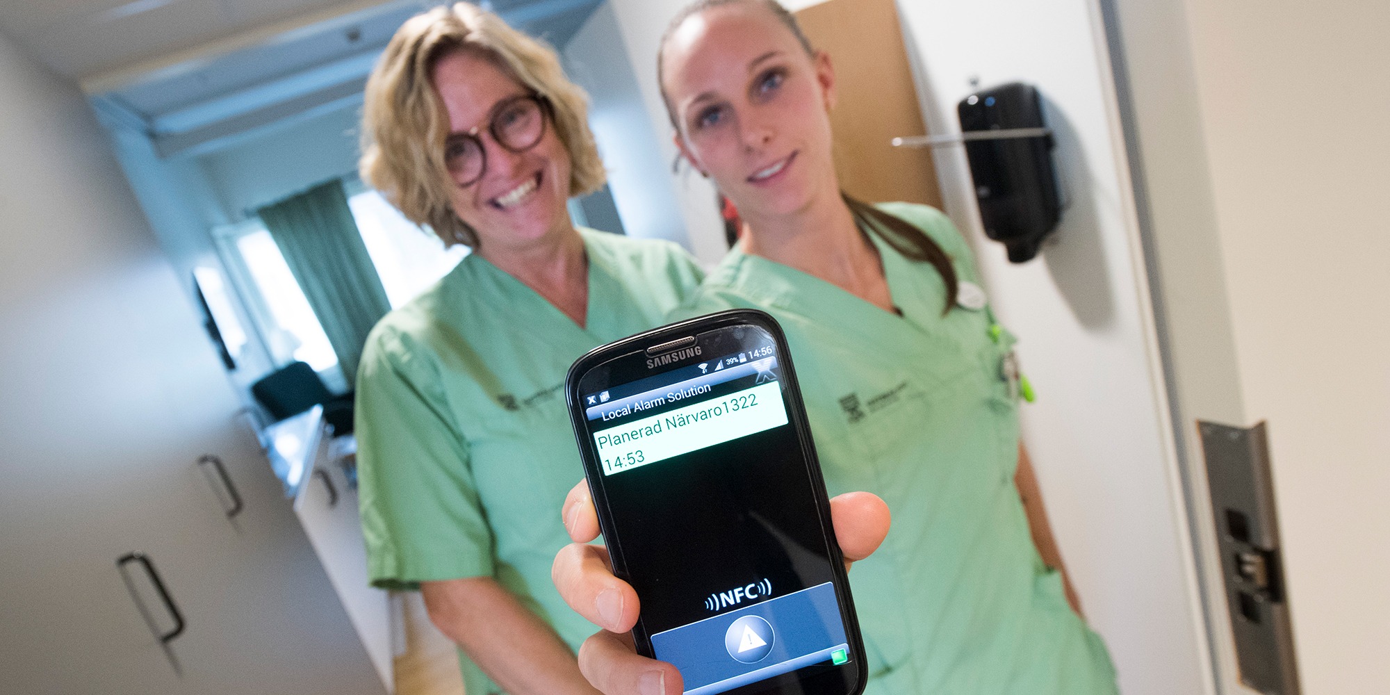 två kvinnor i gröna sjukvårdskläder står tätt intill varandra i en korridor, och håller fram en mobil med texten "Planerad närvaro".
