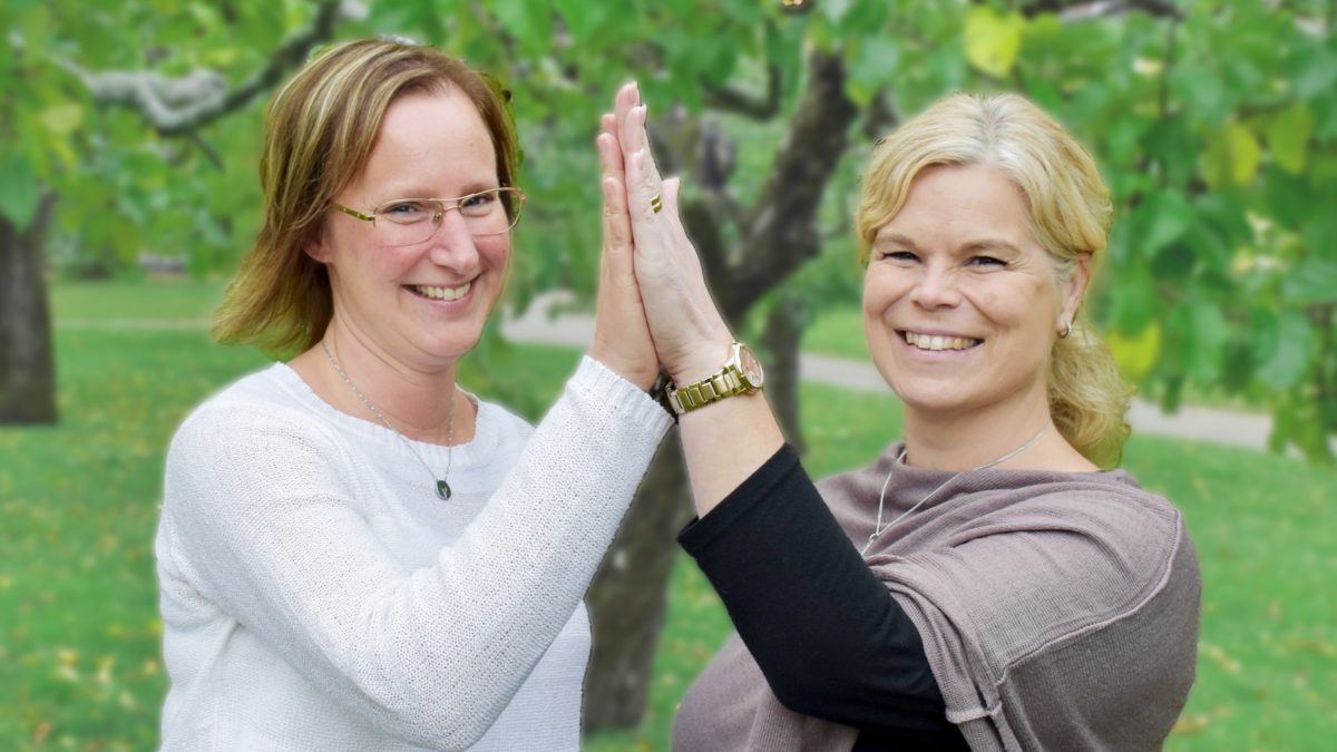 susanne persson och sofia johansson står på en gräsmatta och gör high five mot varandra. de är glada. susanne persson har en vit tröja, sofia johansson har en grå tröja.