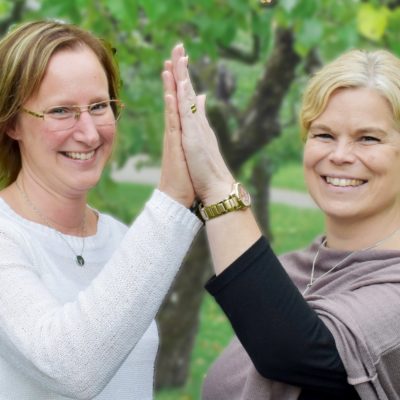 susanne persson och sofia johansson står på en gräsmatta och gör high five mot varandra. de är glada. susanne persson har en vit tröja, sofia johansson har en grå tröja.