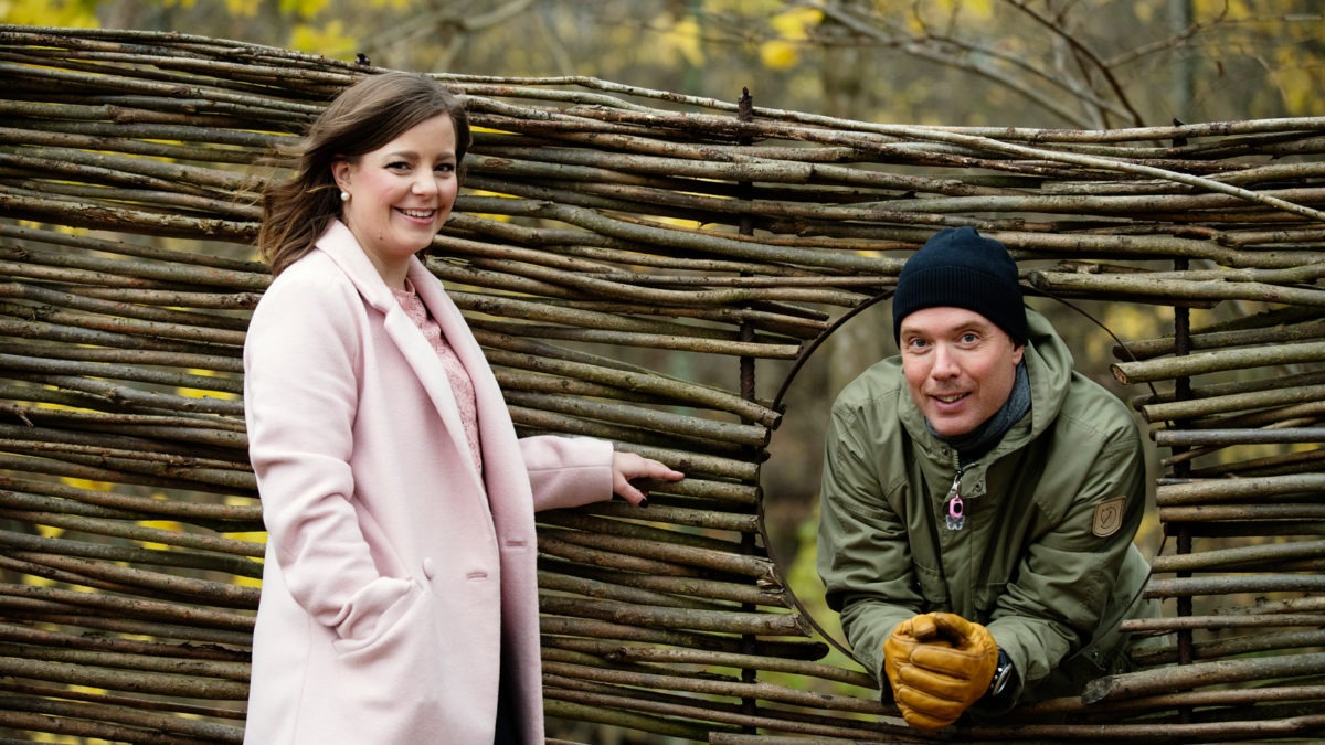 porträtt på Linda Corin, i rosa kappa, och Martin Levin, i grön jacka och svart mössa, som tittar ut genom ett hål i ett staket. Båda ser glada ut. De har båda jobbat med en medarbetarenkät i Västra Götaland.
