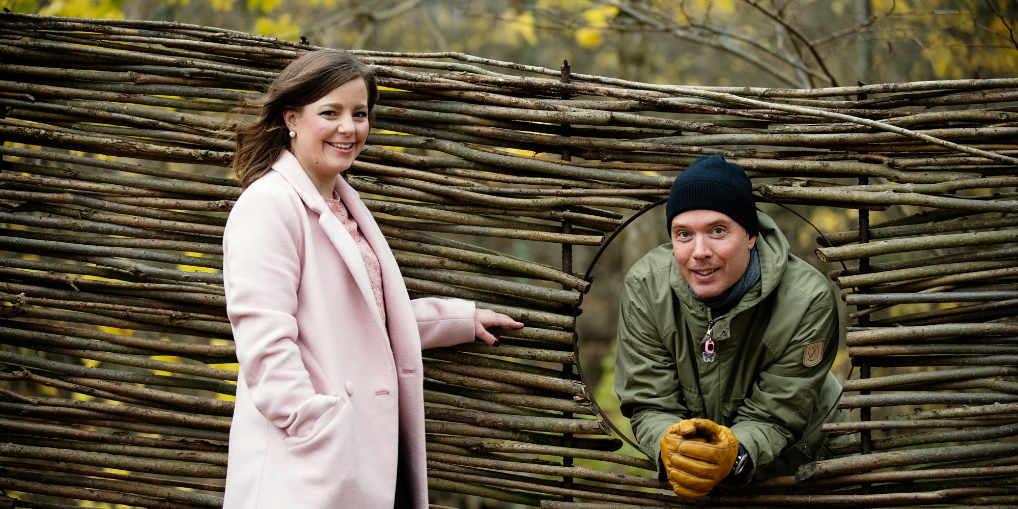 porträtt på Linda Corin, i rosa kappa, och Martin Levin, i grön jacka och svart mössa, som tittar ut genom ett hål i ett staket. Båda ser glada ut. De har båda jobbat med en medarbetarenkät i Västra Götaland.