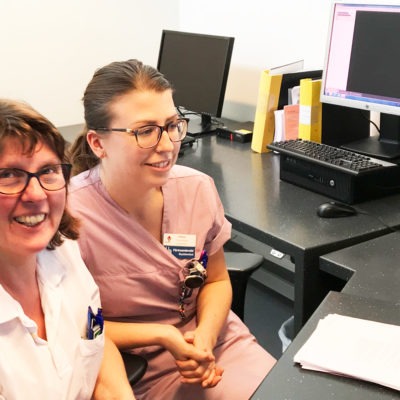 Två kvinnor i vårdkläder vid ett skrivbord bakom en disk på sjukvårdsmottagning. Fotot taget snett uppifrån.