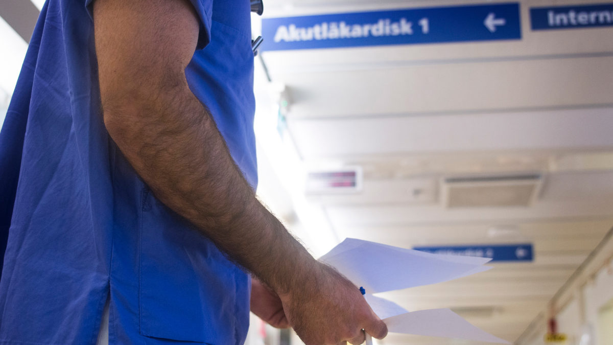 en sjukhuskorridor med skylt i taket som hänvisar till aktuläkardisk. En person i blå vårdkläder står i förgrunden, bara bålen syns. Artikel om skiftarbete.