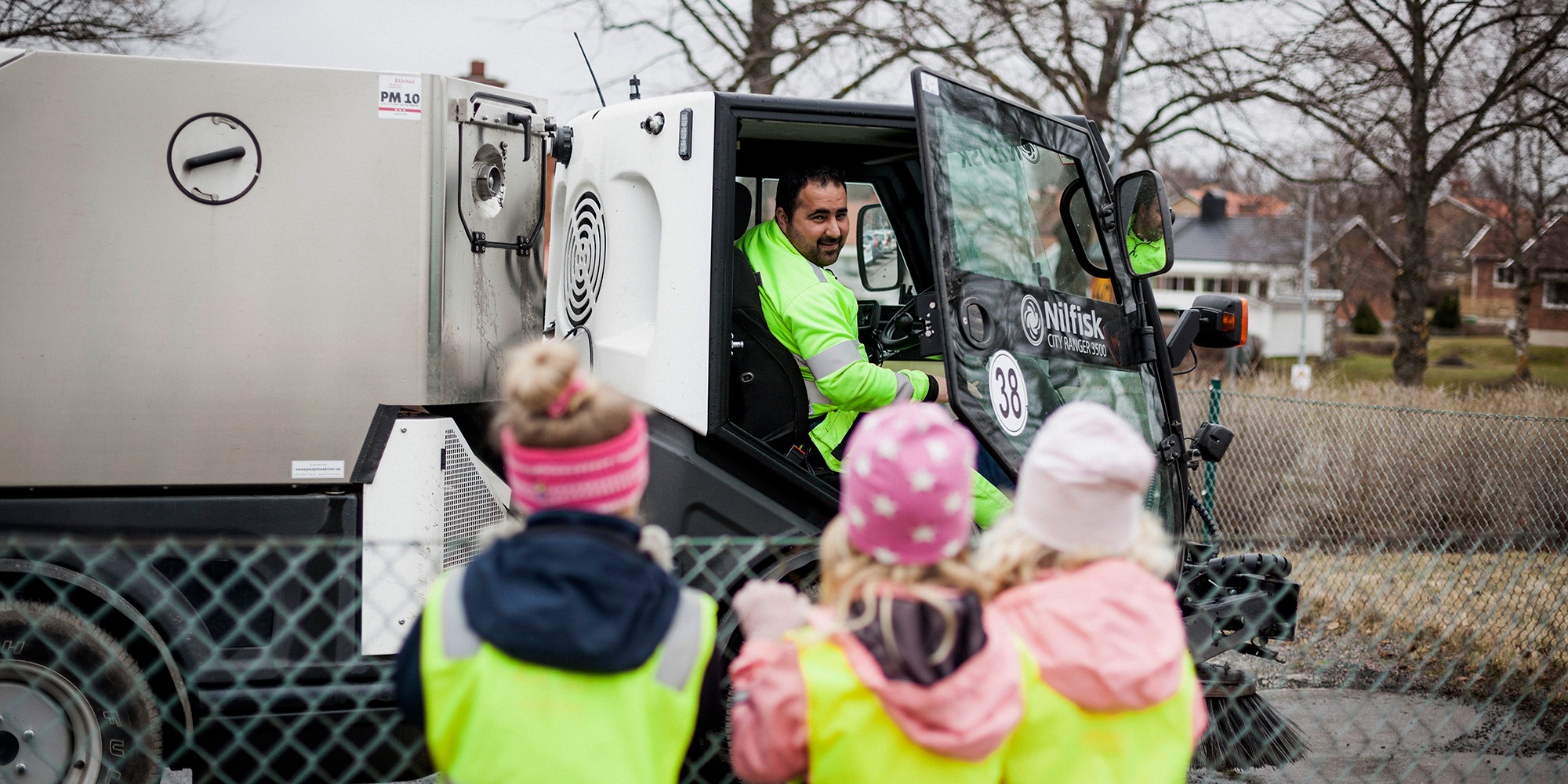 förskolebarn i förgrunden utomhus vid ett staket, sedda bakifrån. bortom dem, i skärpa, en städmaskin med en man i maskinen som öppnat dörren och leende tittar på barnen.