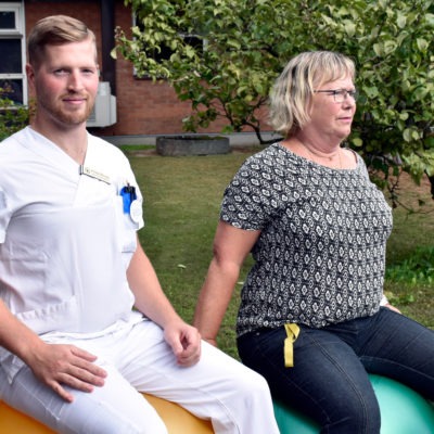 Fysioterapeut och sekreterare sitter på varsin pilatesboll i trädgård utanför vårdcentral, återhämtning i arbetet.