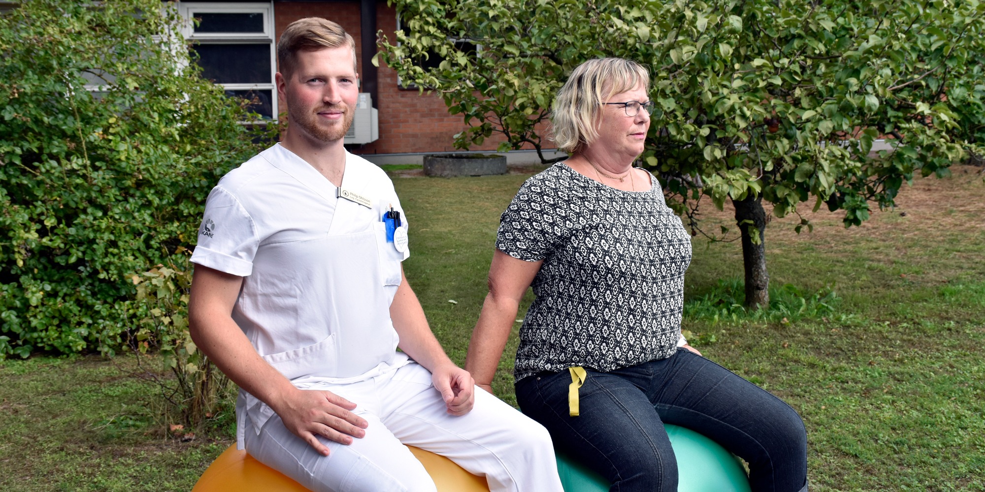 Fysioterapeut och sekreterare sitter på varsin pilatesboll i trädgård utanför vårdcentral, återhämtning i arbetet.