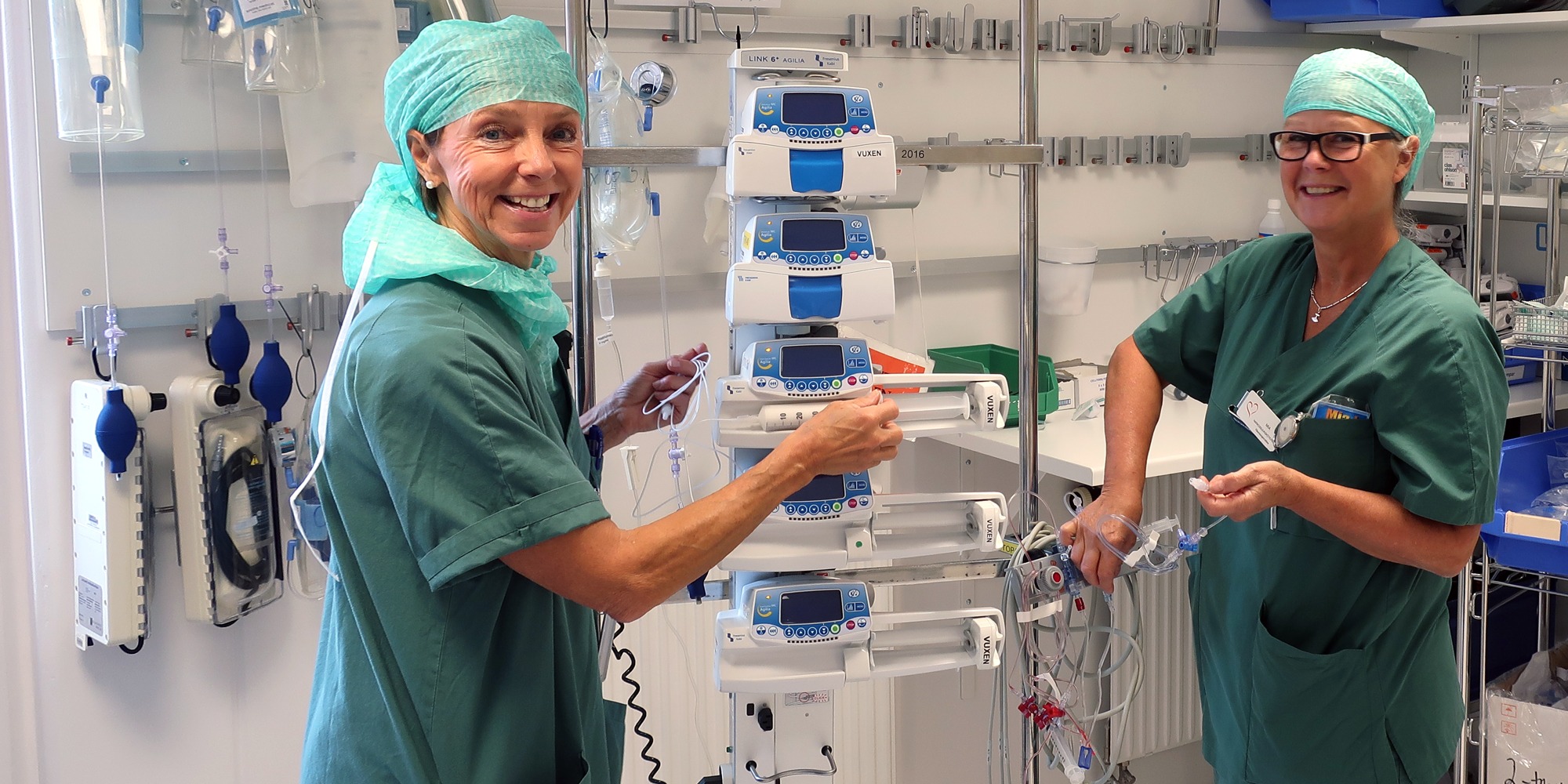 Marika Hasselby Ingmanson och Mia Jensen, i gröna operationskläder, står mitt emot varandra och arbetar med n uppsättning läkemedelspumpar i ett ställ.