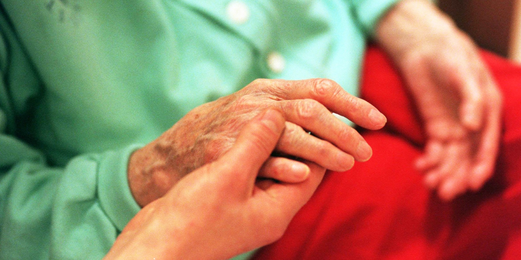 Händer mot ett rött täcke i en säng. En yngre person håller i en äldre persons ena hand.