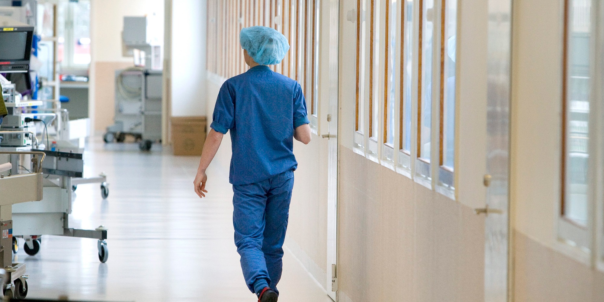 sjukhuskorridor där en person går bort från kameran, klädd i blå operationskläder. Tema vinterkräksjuka.