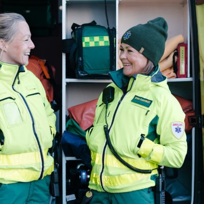 två kvinnliga ambulansförare står mitt emot varandra utanför en ambulans. De ler mot varandra. I bakgrunden syns den öppna ambulansen full med utrustning.