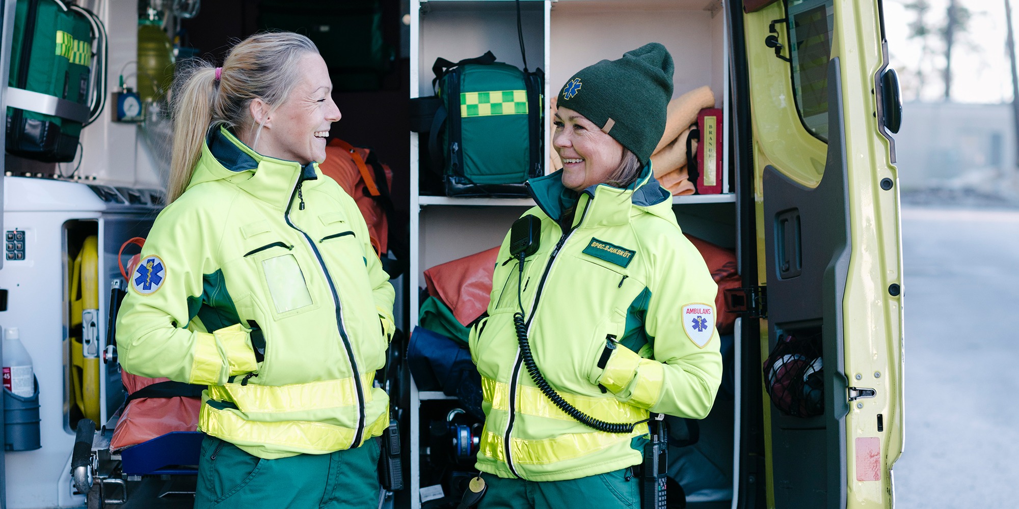 två kvinnliga ambulansförare står mitt emot varandra utanför en ambulans. De ler mot varandra. I bakgrunden syns den öppna ambulansen full med utrustning.