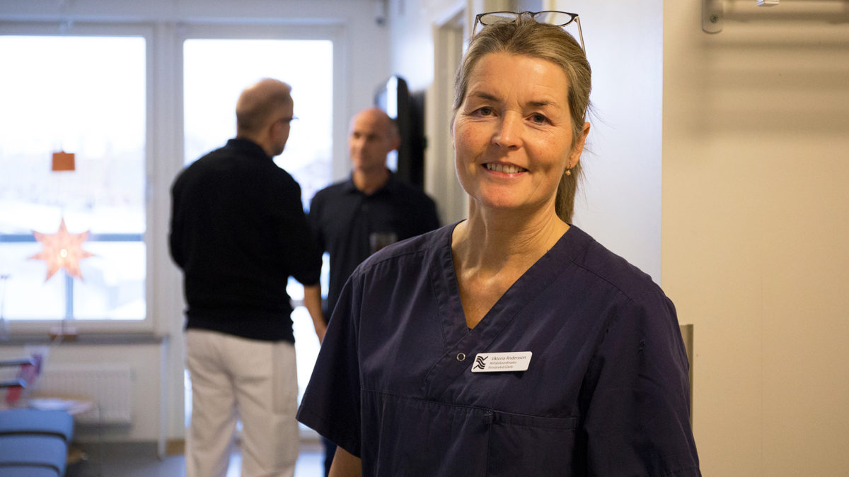 Porträtt på Viktoria Andersson, rehabkoordinator på Gävle Strands hälsocentral. Viktoria har en märk sjukhusblus på sig och står i en korridor på hälsocentralen, med ett par personer i bakgrunden.