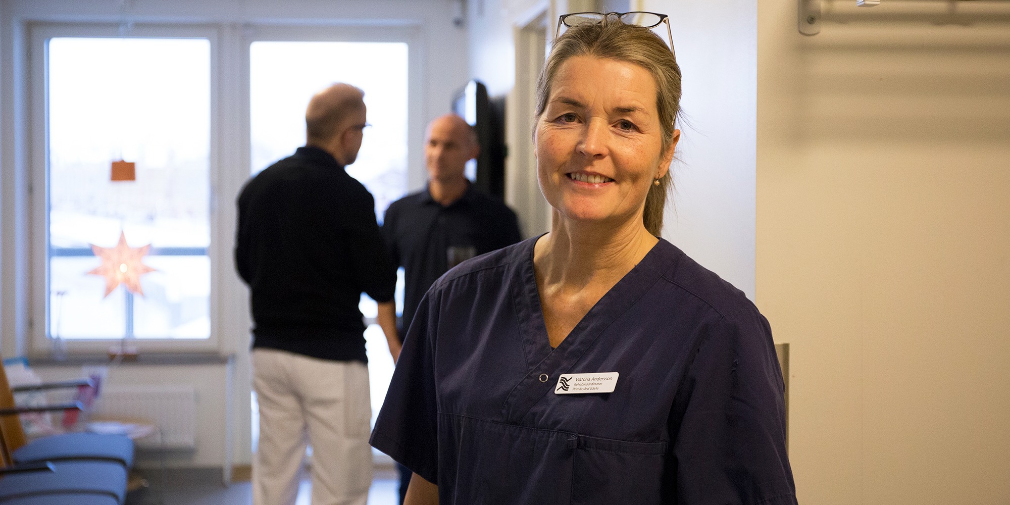 Porträtt på Viktoria Andersson, rehabkoordinator på Gävle Strands hälsocentral. Viktoria har en märk sjukhusblus på sig och står i en korridor på hälsocentralen, med ett par personer i bakgrunden.