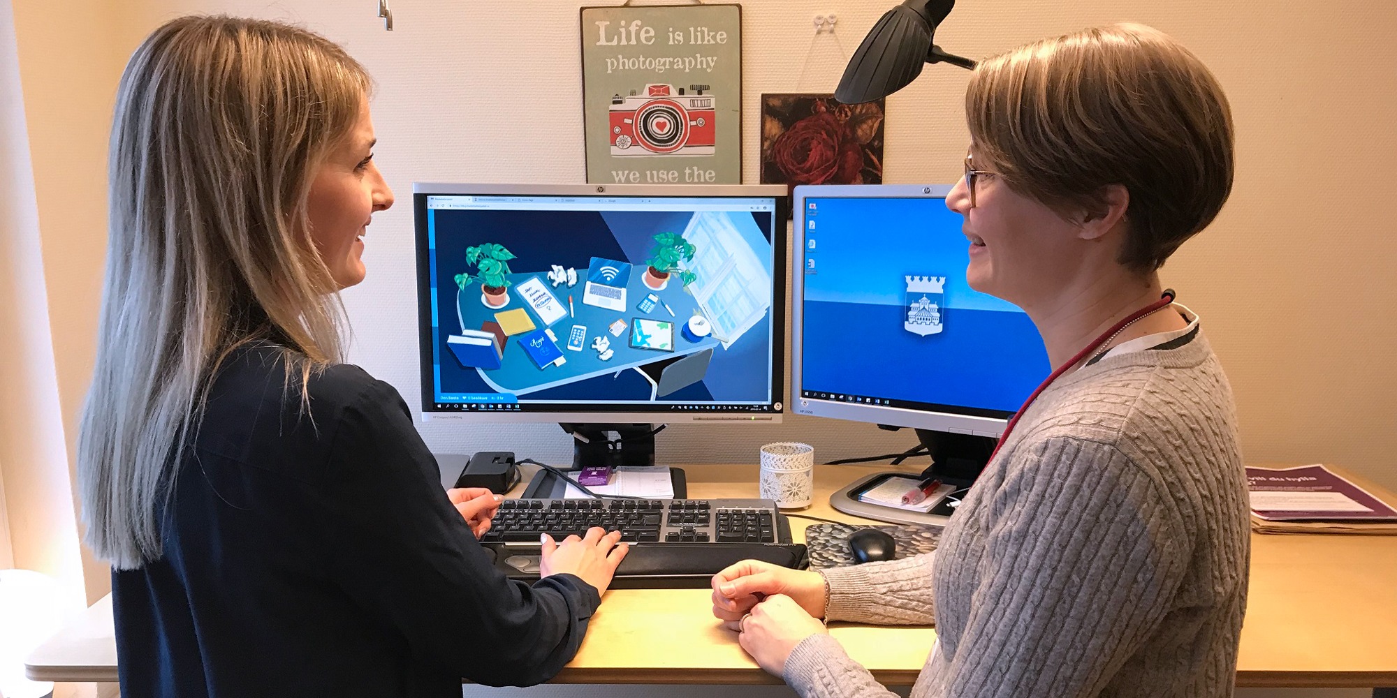 Två kvinnor står vid ett skrivbord med datorskärmar och spelar Helsingborgs stads datorspel om medarbetarskap, tema gamification.