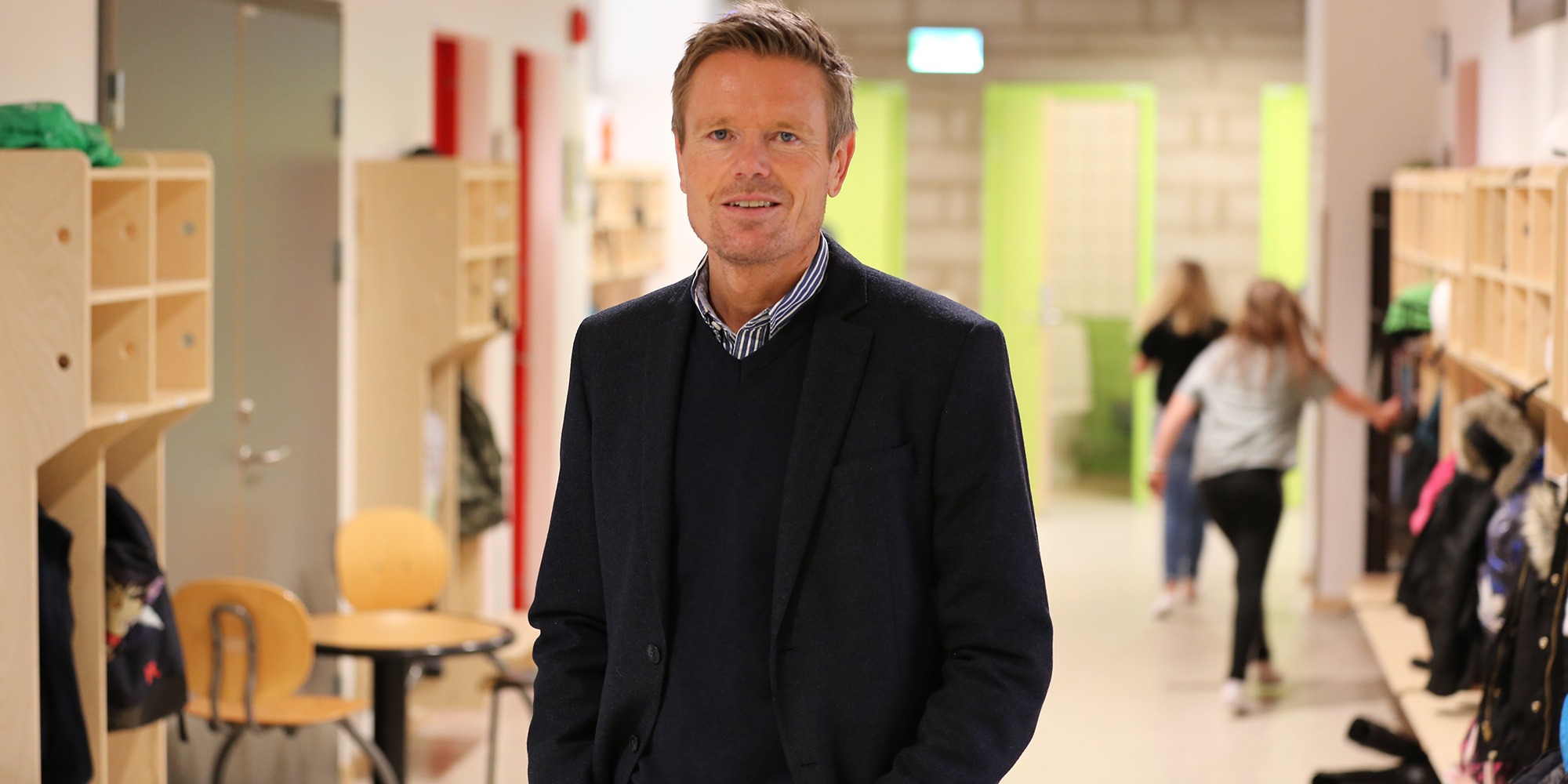 porträtt av Jonas Hällström, rektor på Fladängskolan i Lomma komun. Bilden tagen i en skolkorridor med springande barn bakom honom.