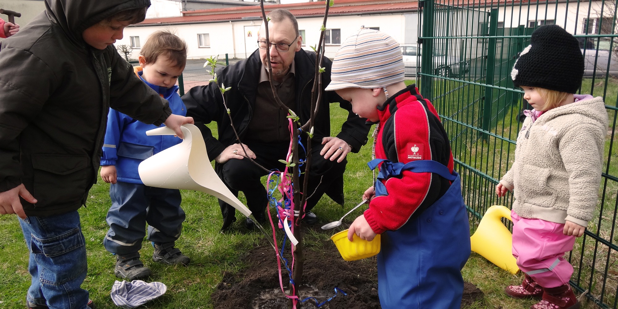 Tre små barn i galonkläder hjälps åt att vattna en planta. i ringen med barnen sitter en vuxen äldre man på huk.