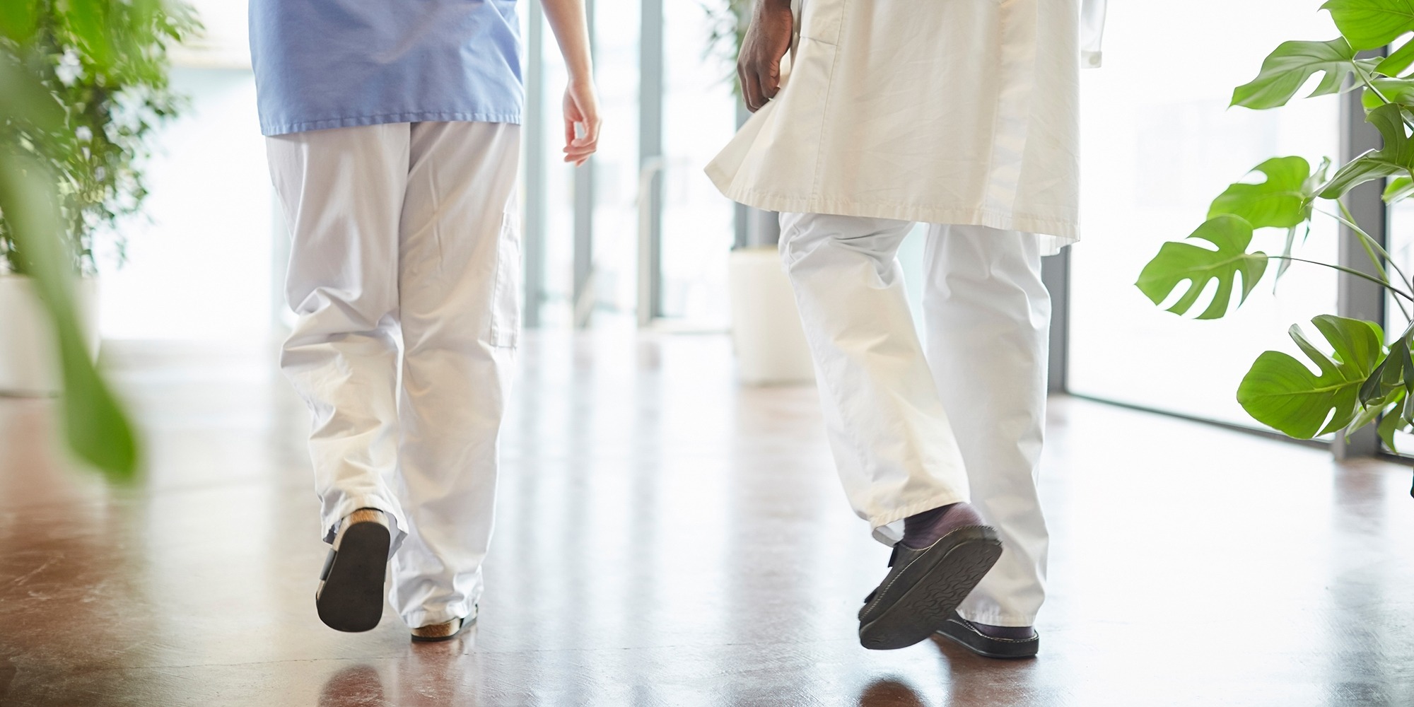 Fallolyckor. Två personer i sjukhuskläder syns bakifrån gå i en ljus sjukhuskorridor med blankt golv.
