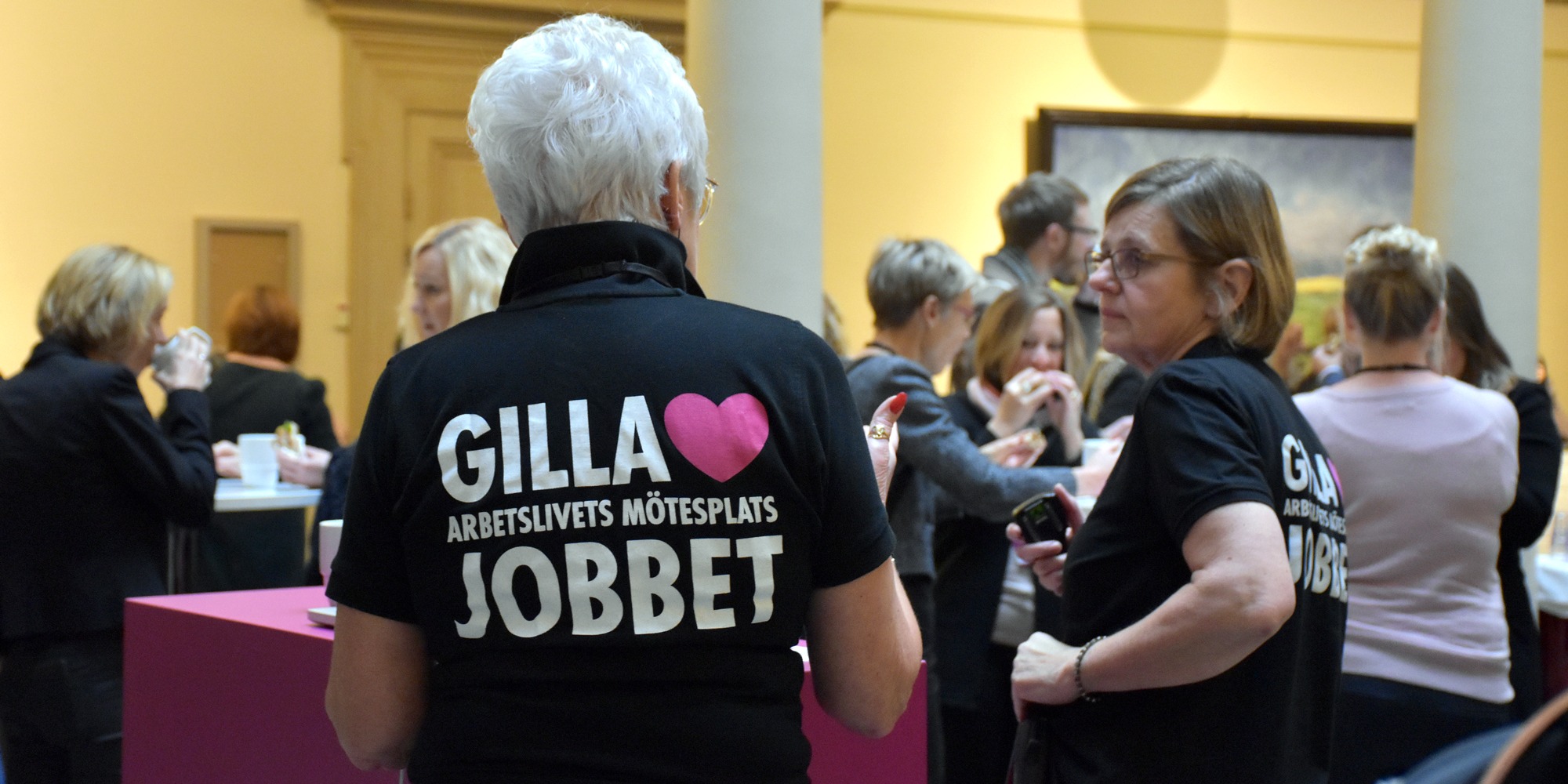 en grupp människor med ryggen mot betraktaren, har på sig svarta tröjor med rosa GillaJobbet-logga på ryggen.