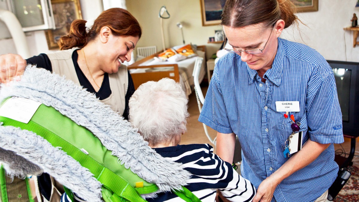 två undersköterskor i arbetskläder hjälper en gammal kvinna att sätta sig i sin stol, i hemmiljö.