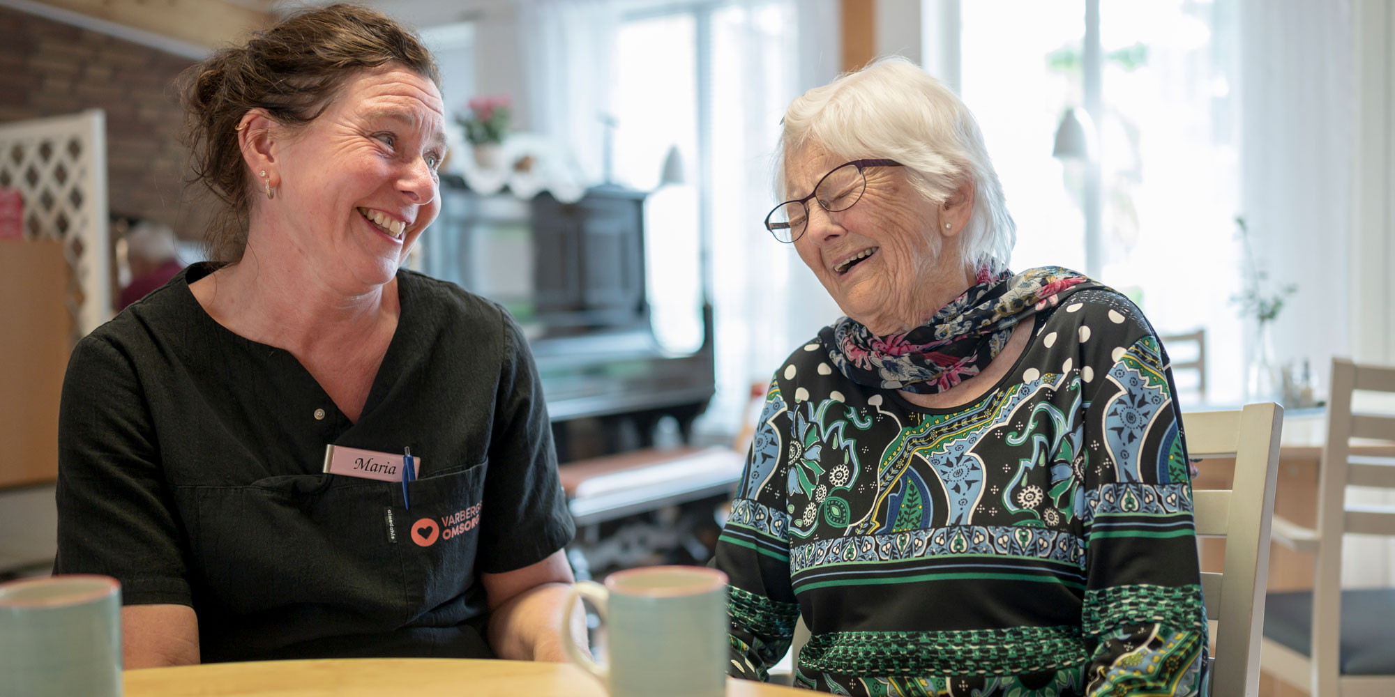 En kvinna i vårdkläder skrattar tillsammans med en äldre kvinna vid ett bord.