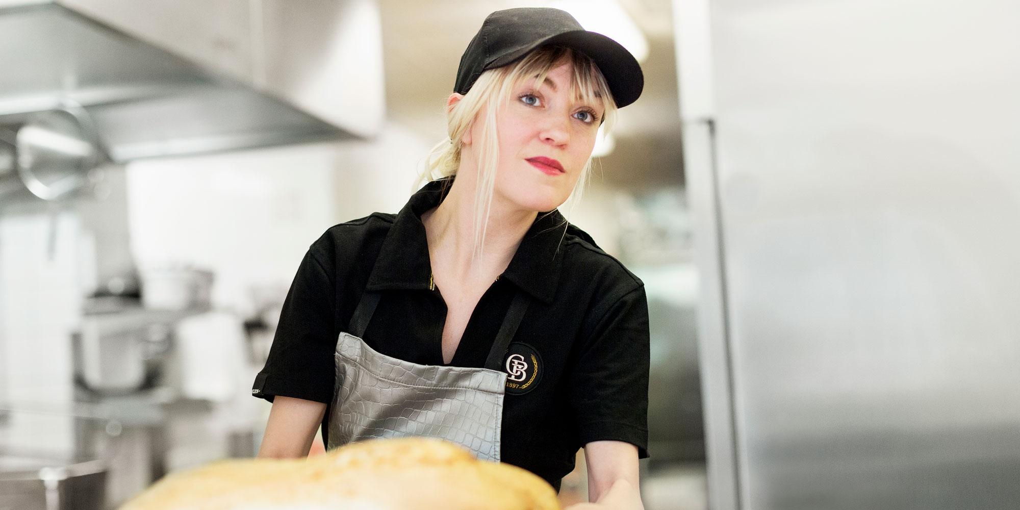 Blond kvinna i arbetsskjorta och keps lyfter ett bröd, i storköksmiljö.
