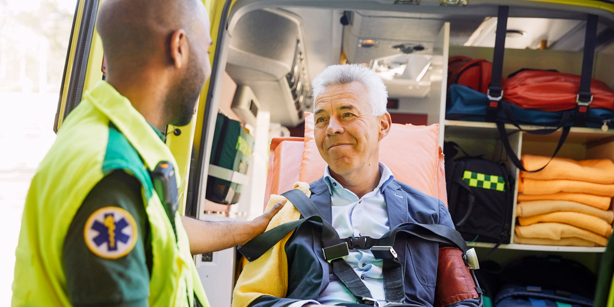 En äldre man sitter på en bår utanför en öppen ambulans. Han ler mot en ambulanssjukvårdare som har ryggen mot kameran.