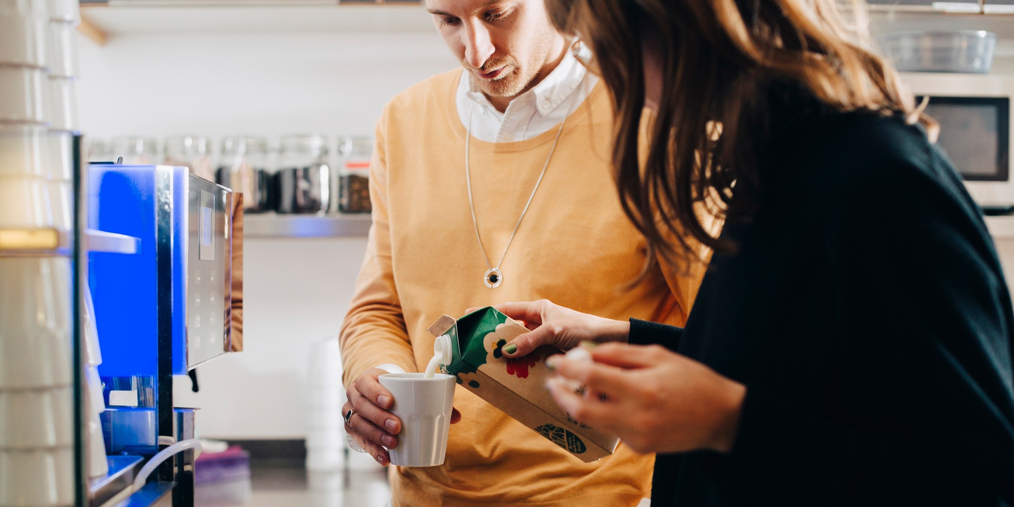 En man och en kvinna står vid en kaffeautomat. Kvinnan häller upp mjölk åt mannen.