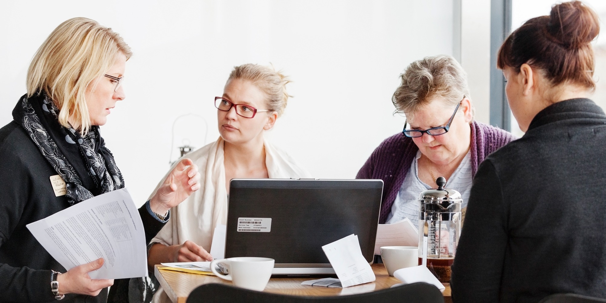 Fyra personer sitter och tittar i olika papper vid ett skrivbord. Öppen laptop och kaffekoppar står på bordet.