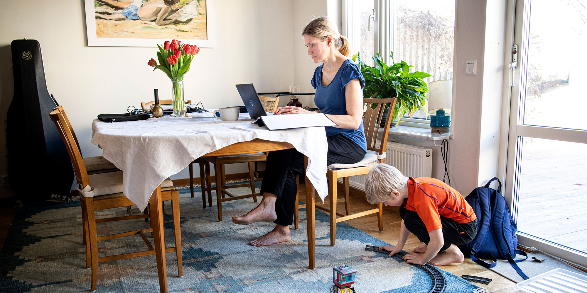 Kvinna som arbetar framför dator vid matsalsbord hemma. BArm leker med tågbana under bordet.