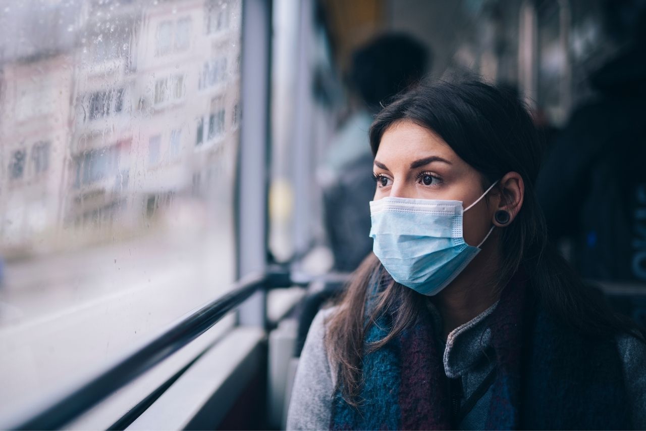 Kvinna med blått munskydd sitter på en buss och tittar ut genom ett regnigt fönster