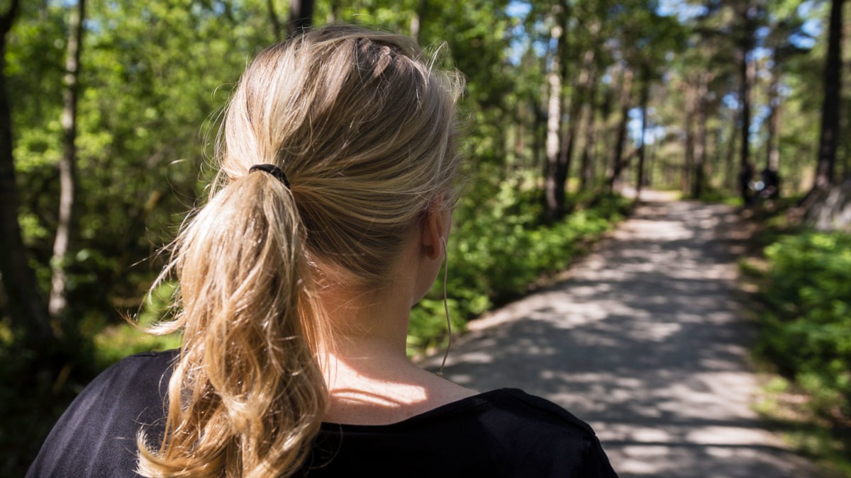 Kvinna sedd bakifrån med blond hästsvans, på en somrig skogsstig.