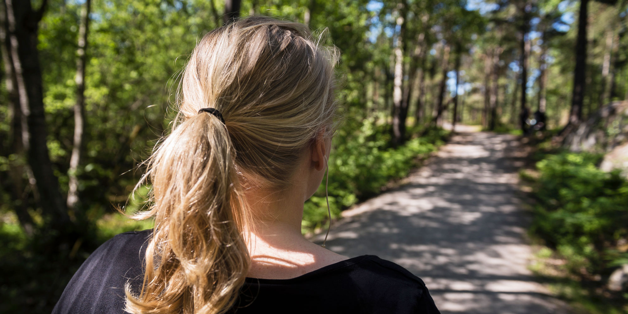 Kvinna sedd bakifrån med blond hästsvans, på en somrig skogsstig.