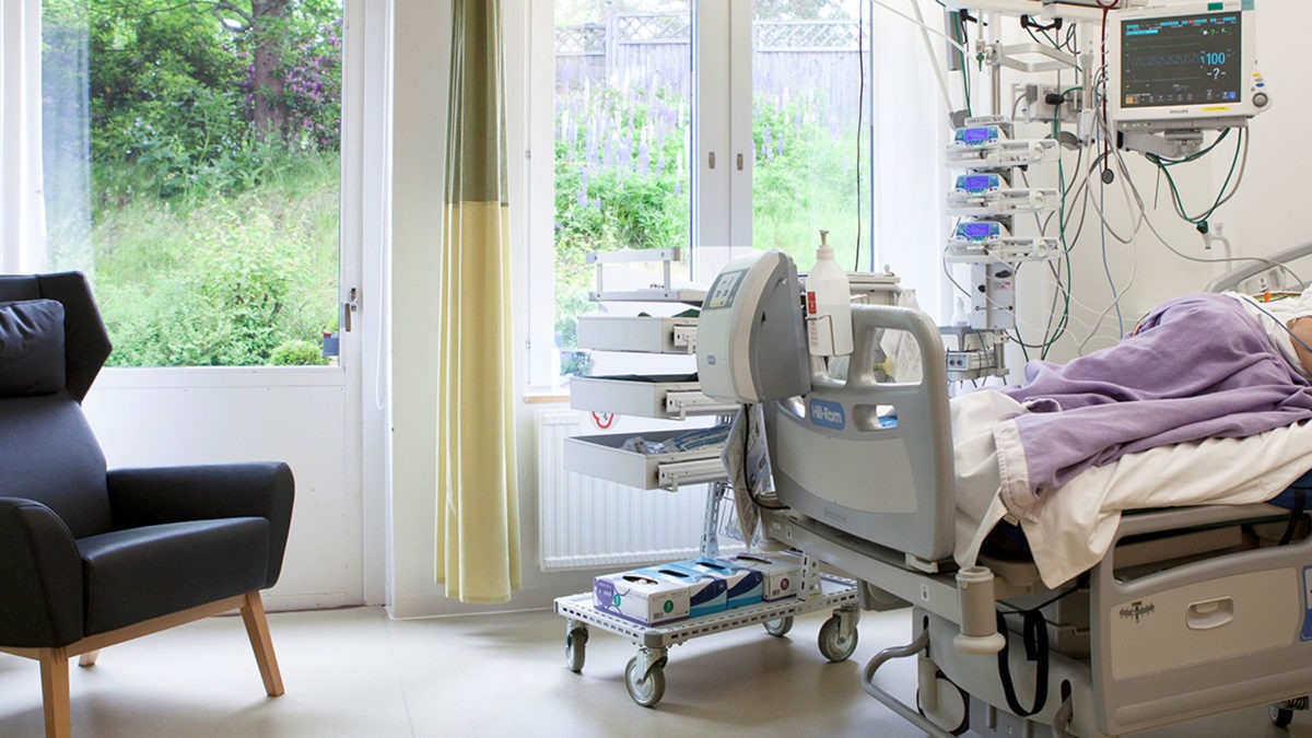 Ett ljust rum på sjukhus avsett för intensivvård, med säng, fåtölj och fönster med skira gardiner.