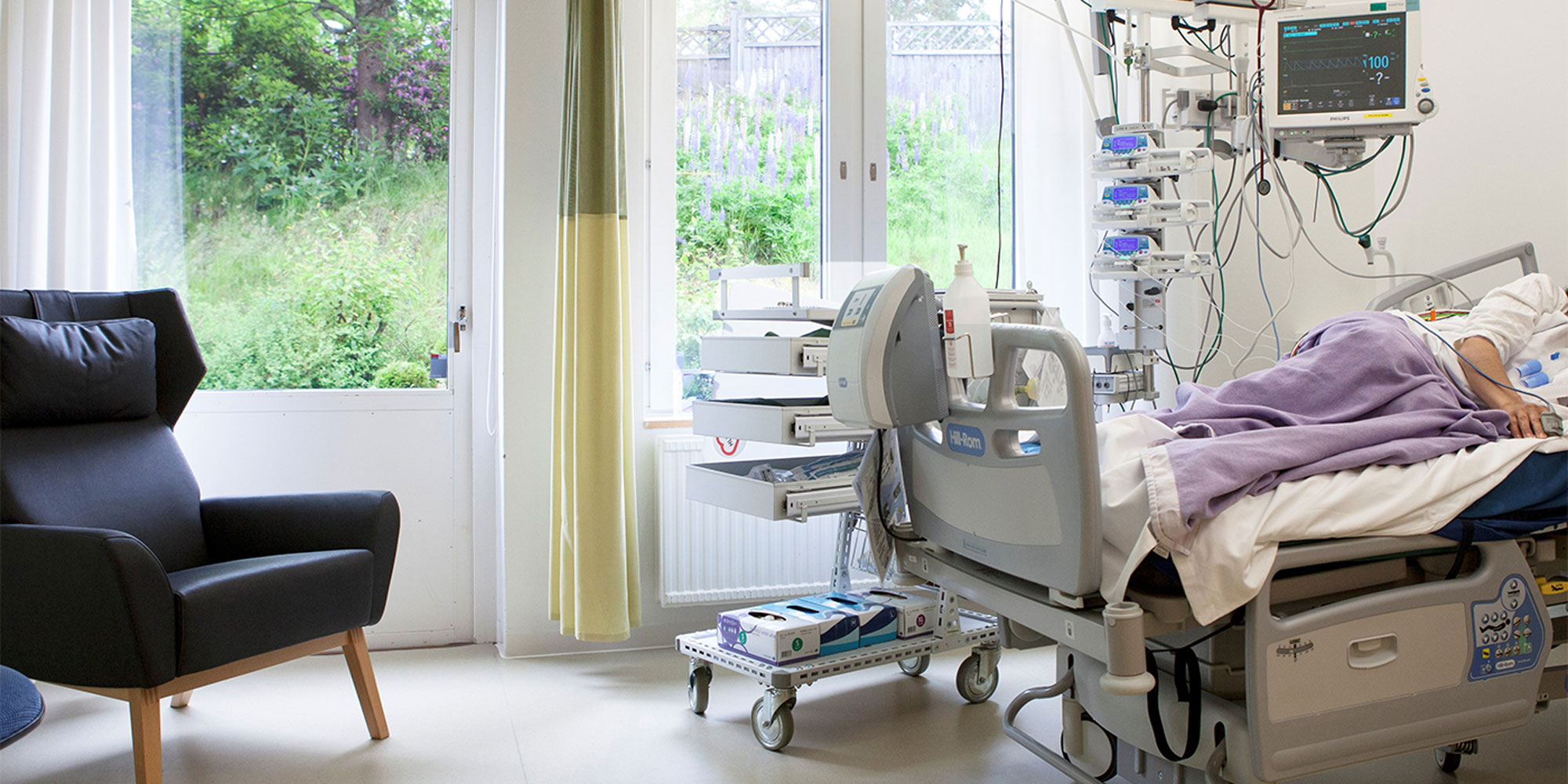 Ett ljust rum på sjukhus avsett för intensivvård, med säng, fåtölj och fönster med skira gardiner.