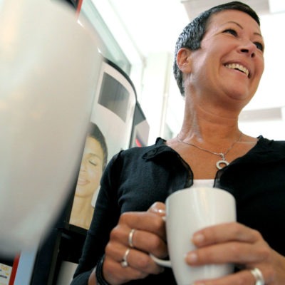 Leende kvinna med kaffemugg. Runt henne flera andra kaffemuggar som hålls av händer.