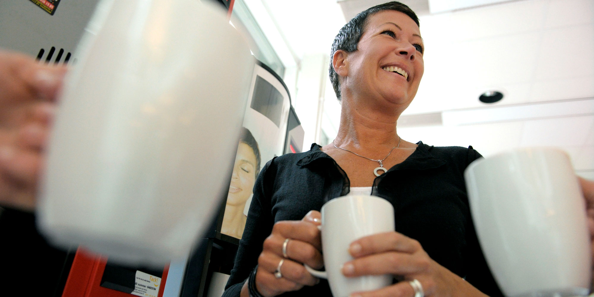 Leende kvinna med kaffemugg. Runt henne flera andra kaffemuggar som hålls av händer.