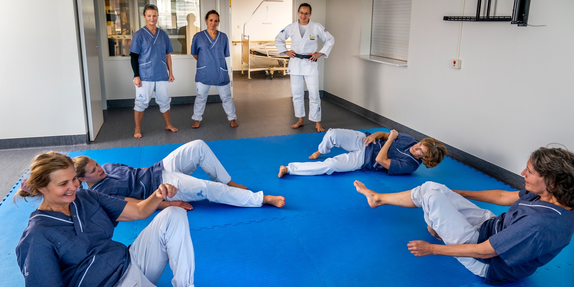 Fyra personer i sjukhuskläder tränar fall på en blå gymnastikmatta. Tre andra personer ser på.